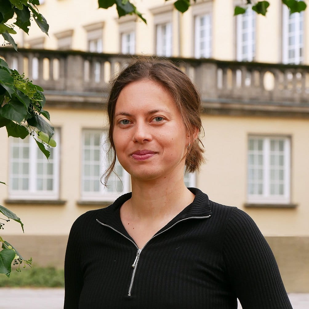 Profilfoto von Nele Becker, im Sonnenschein vor grün bepflanztem Hintergrund.